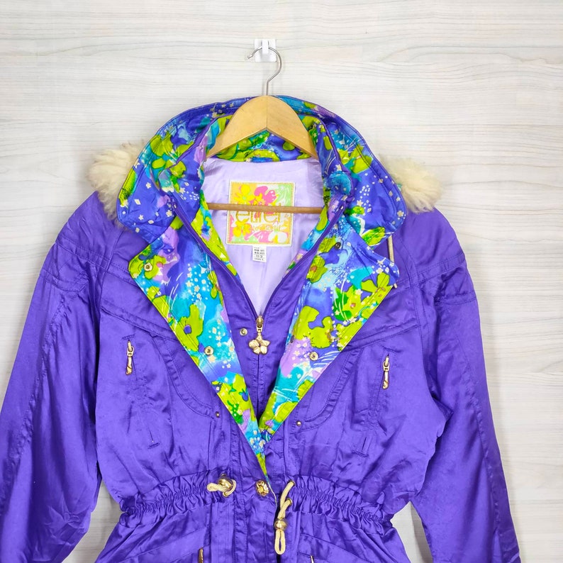 May include: A purple ski jacket with a floral lining and a fur-trimmed hood. The jacket has a zipper closure, a drawstring waist, and gold-toned hardware. The label on the jacket reads "Ellen Tracy".