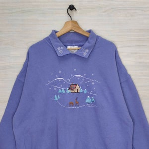 May include: A purple sweatshirt with a winter scene embroidered on the front. The scene includes a small house, trees, and two deer. The collar is trimmed with white snowflakes.