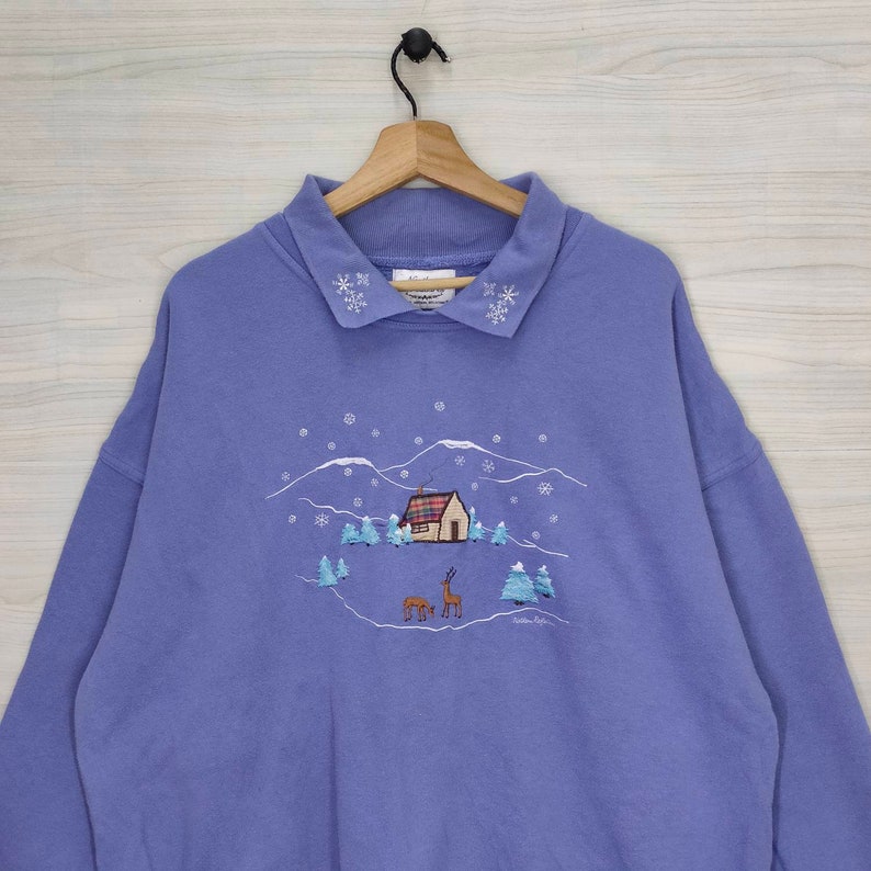 May include: A purple sweatshirt with a winter scene embroidered on the front. The scene includes a small house, trees, and two deer. The collar is trimmed with white snowflakes.