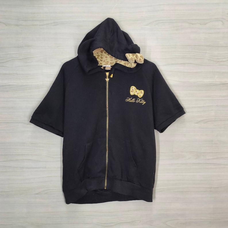 May include: Black hooded zip-up sweatshirt with short sleeves. Features a gold zipper, a gold bow and "Hello Kitty" embroidered on the front. The hood lining has a tan and brown animal print.
