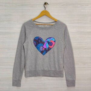 Puede incluir: Una sudadera gris con un gran estampado en forma de corazón con un diseño floral colorido. El corazón está contorneado en azul.