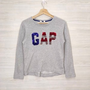 May include: A gray long-sleeved sweatshirt with the word "GAP" in red, white, and blue sequins.