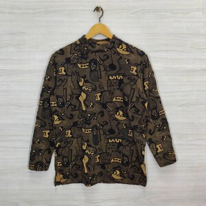 Op de afbeelding: Een shirt met lange mouwen en een hoge kraag, met een herhaald patroon van zwarte en bruine abstracte vormen. Het shirt hangt aan een houten hanger tegen een lichte achtergrond.