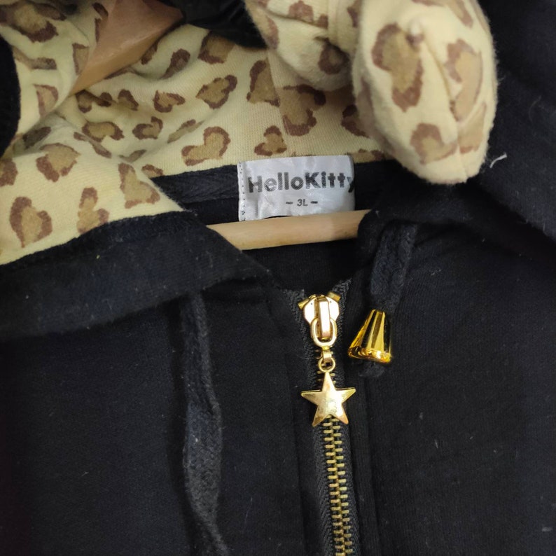 May include: Black zip-up hoodie with a leopard print lining and hood detail. The zipper has a gold star charm. The label reads "Hello Kitty - 3L".
