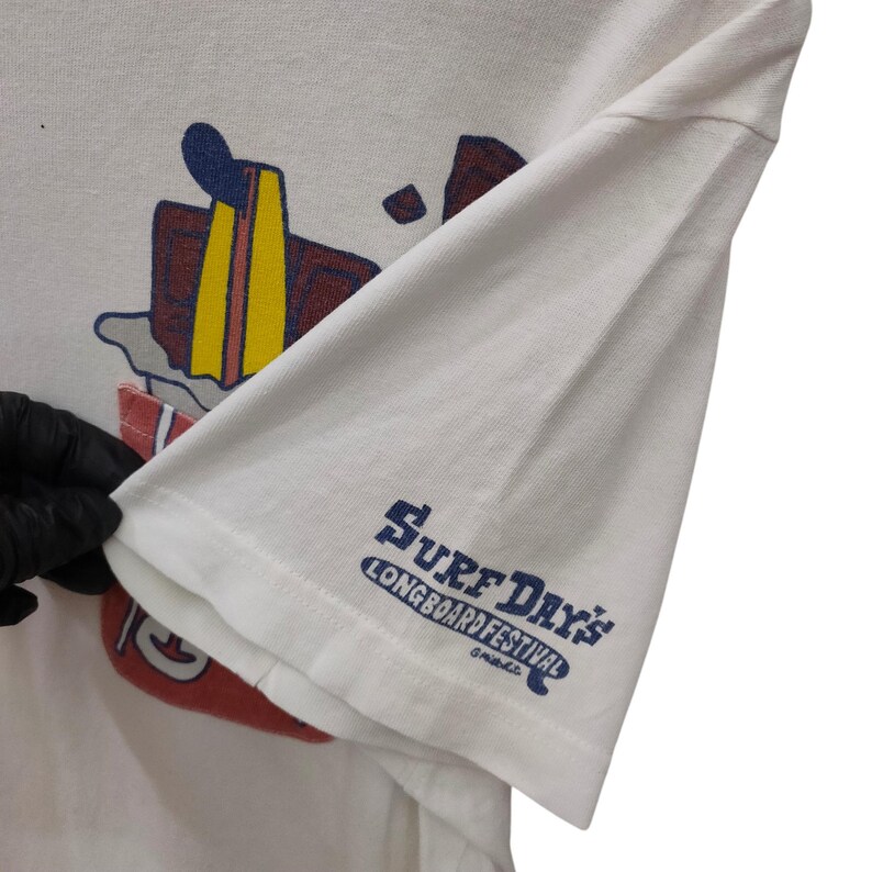 May include: White t-shirt with a colorful graphic of a surfboard and other surf-related imagery. The sleeve features the text "Surf Day's Longboard Festival" in blue. The shirt is made of a soft, likely cotton material.