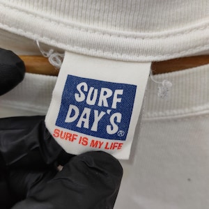 May include: Close-up of a white t-shirt tag with the text "SURF DAY'S" in white letters on a blue background. Below the brand name, the text "SURF IS MY LIFE" is printed in red. The tag is attached to a white t-shirt.