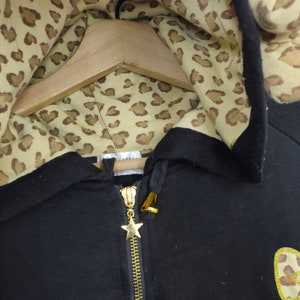 May include: Black zip-up hoodie with a gold zipper and star charm. The hood lining features a tan and brown leopard print pattern. The hoodie is hanging on a wooden hanger.