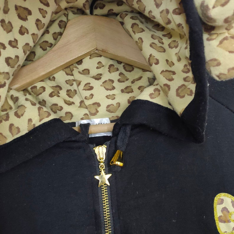 May include: Black zip-up hoodie with a gold zipper and star charm. The hood lining features a tan and brown leopard print pattern. The hoodie is hanging on a wooden hanger.