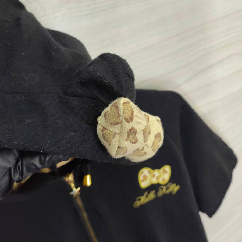 May include: Black hooded zip-up sweatshirt with a gold zipper and a bow-shaped accent on the hood. The bow is tan with brown spots. The sweatshirt has the words "Hello Kitty" embroidered in gold.