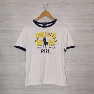 May include: White t-shirt with a blue and yellow Polo Ralph Lauren logo. The logo features a polo player on a horse and the text "Polo 1967 Nine Sixty Seven" and "P.R.I.". The shirt has a blue and white striped collar and cuffs.