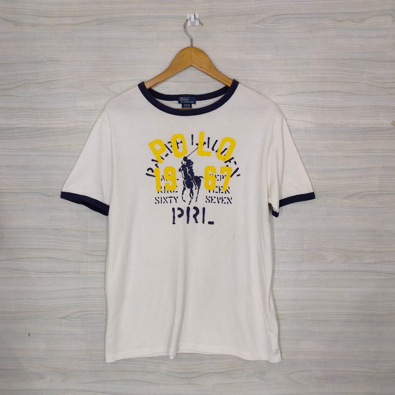 May include: White t-shirt with a blue and yellow Polo Ralph Lauren logo. The logo features a polo player on a horse and the text "Polo 1967 Nine Sixty Seven" and "P.R.I.". The shirt has a blue and white striped collar and cuffs.
