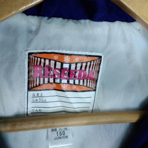 May include: A white label with the word "RESEDA" in red and black letters. The label has a red and white striped border and snowflakes. The label also has a white tag with Japanese text and a white tag with the text "150 JUNIOR" in black letters.