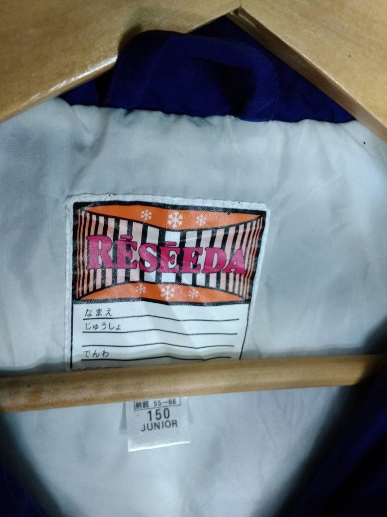 May include: A white label with the word "RESEDA" in red and black letters. The label has a red and white striped border and snowflakes. The label also has a white tag with Japanese text and a white tag with the text "150 JUNIOR" in black letters.