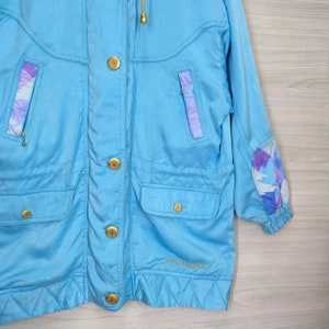May include: A light blue jacket with a gold zipper and buttons. The jacket has a hood and a  "Courr&egrave;ges" logo on the bottom. The sleeves have a purple and white geometric pattern.