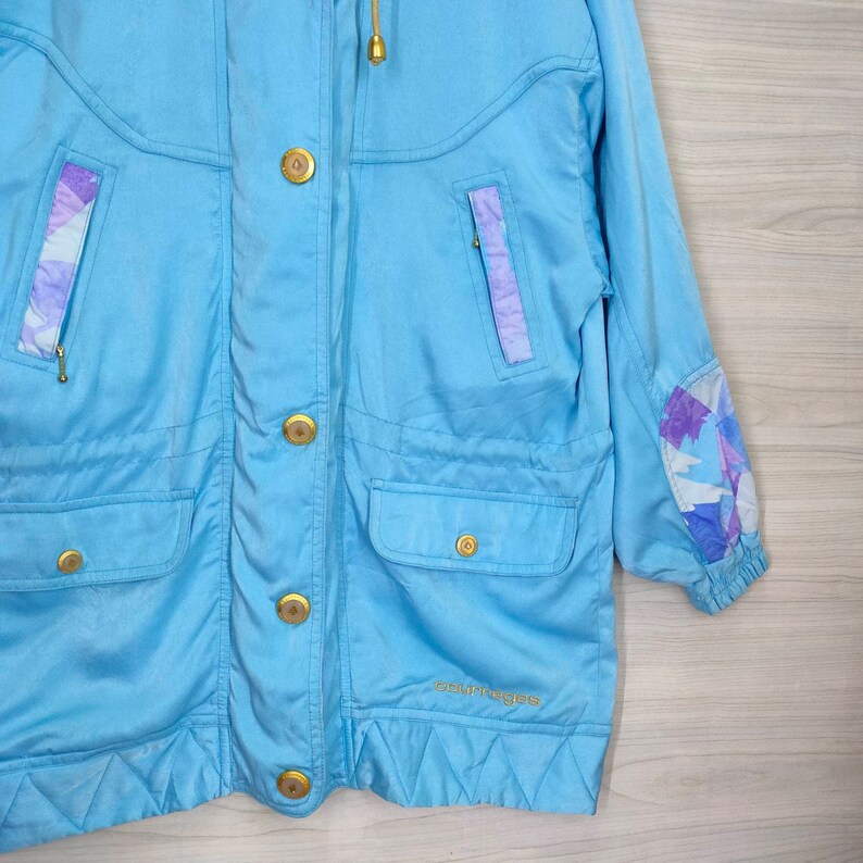 May include: A light blue jacket with a gold zipper and buttons. The jacket has a hood and a  "Courr&egrave;ges" logo on the bottom. The sleeves have a purple and white geometric pattern.