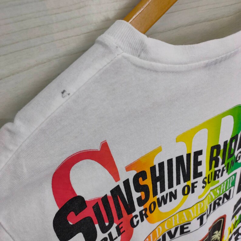 Puede incluir: Camiseta blanca con un gr&aacute;fico colorido que dice "Sunshine Ride" en letras grandes y estilizadas. El dise&ntilde;o incluye texto rojo, amarillo, verde y negro, con texto adicional que indica un campeonato de surf. La camiseta se muestra en una percha de madera.