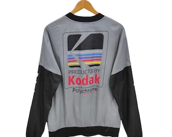 Puede incluir: Chaqueta cortavientos vintage Kodak gris y negra. La chaqueta presenta el logotipo de Kodak en la espalda con las palabras "PRODUCTS BY Kodak Polychrome". Las mangas son negras y el cuerpo gris.
