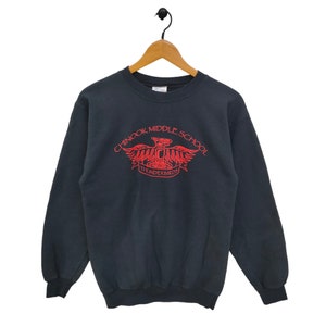 Pode incluir: Camisola preta de gola redonda com o texto "Chinook Middle School" em vermelho, um gráfico de águia vermelha e a palavra "Thunderbirds". A camisola está pendurada num cabide de madeira.