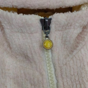 May include: Close-up of a zipper on a pink and white fleece jacket. The zipper pull has a yellow logo that reads "Courr&egrave;ges Sport Futur".