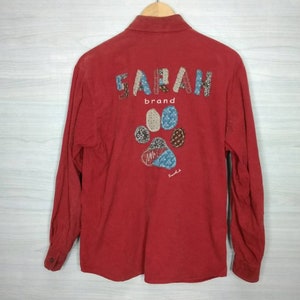 May include: Red long-sleeved button-up shirt with a colourful patchwork design of the word "SARAH" and a paw print. The text "brand" is below the word "SARAH".