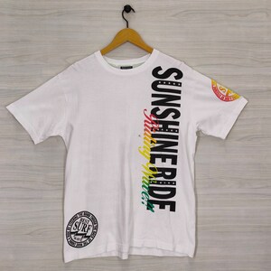 Puede incluir: Camiseta blanca con las palabras "SUNSHINE RIDE" en texto vertical negro y una escritura colorida "making waves". La camiseta tambi&eacute;n presenta un gr&aacute;fico circular con las palabras "BEST SURF" y un peque&ntilde;o logotipo en la manga.