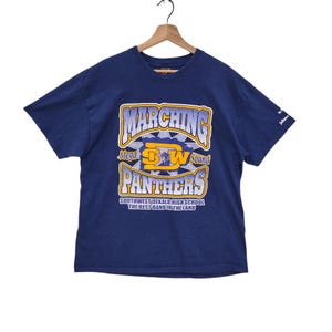 Pode incluir: Uma camiseta azul com um gráfico amarelo e roxo de um logotipo de banda de marcha. O logotipo diz "Marching Mega DW Sound Panthers" e "Southwest Dekalb High School The Best Band In The Land".