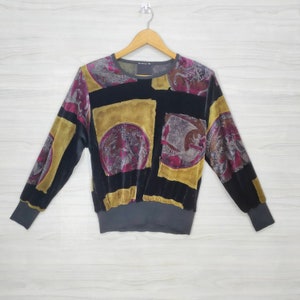 May include: A black long-sleeved shirt with a colourful, abstract print featuring gold, purple, and red accents. The print includes circular designs with figures and a geometric pattern.