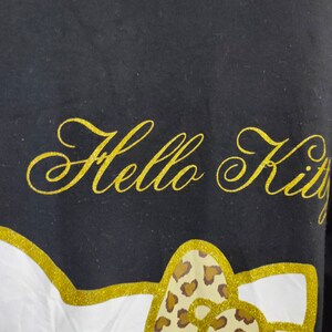 May include: Black t-shirt with the words "Hello Kitty" in gold glitter script. The design includes a gold glitter outline of a bow with a leopard print pattern.