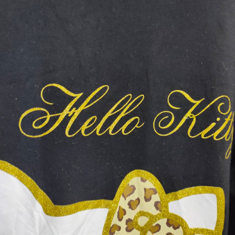 May include: Black t-shirt with the words "Hello Kitty" in gold glitter script. The design includes a gold glitter outline of a bow with a leopard print pattern.