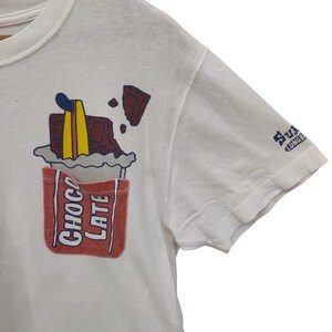 May include: White t-shirt with a whimsical chocolate bar graphic. The design features a red pocket with the word "CHOCOLATE" and a melting chocolate bar with blue, yellow, and brown accents. The sleeve has a small blue logo.