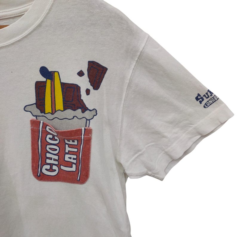 May include: White t-shirt with a whimsical chocolate bar graphic. The design features a red pocket with the word "CHOCOLATE" and a melting chocolate bar with blue, yellow, and brown accents. The sleeve has a small blue logo.