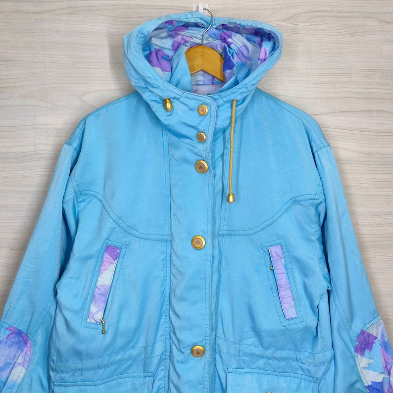 May include: A light blue jacket with a hood and a floral pattern lining. The jacket has a zipper closure and multiple pockets. The jacket has gold buttons.