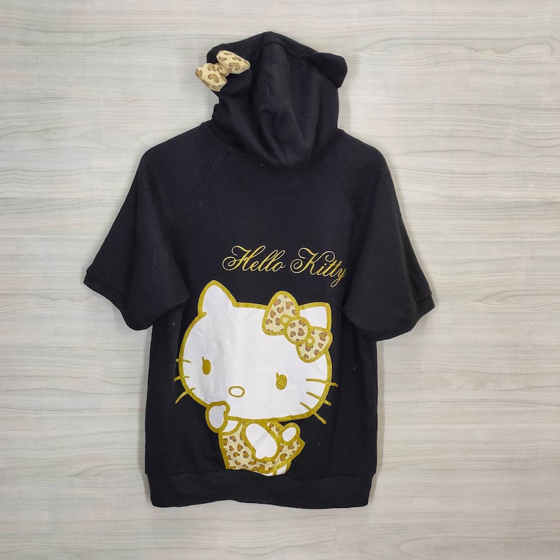 May include: Black short-sleeved hooded sweatshirt featuring a Hello Kitty graphic. The design includes a white Hello Kitty face with a gold bow and accents. The words "Hello Kitty" are written in gold script above the graphic. The hood has ears and bows.