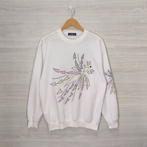 May include: White sweatshirt with a colorful abstract design featuring arrows and geometric shapes. The design is centered on the front of the sweatshirt and extends onto the sleeves.