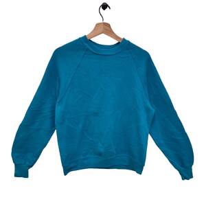 Op de afbeelding: Een tealkleurige sweatshirt met lange mouwen en een ronde hals, hangend aan een houten kleerhanger. De raglanmouwen lopen van de halslijn tot de oksel. De sweatshirt is gemaakt van zacht materiaal, mogelijk katoen.