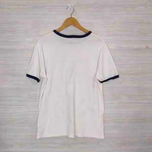 May include: White t-shirt with a navy blue ringer neckline and sleeve trim. The shirt is hanging on a wooden hanger against a woodgrain background.