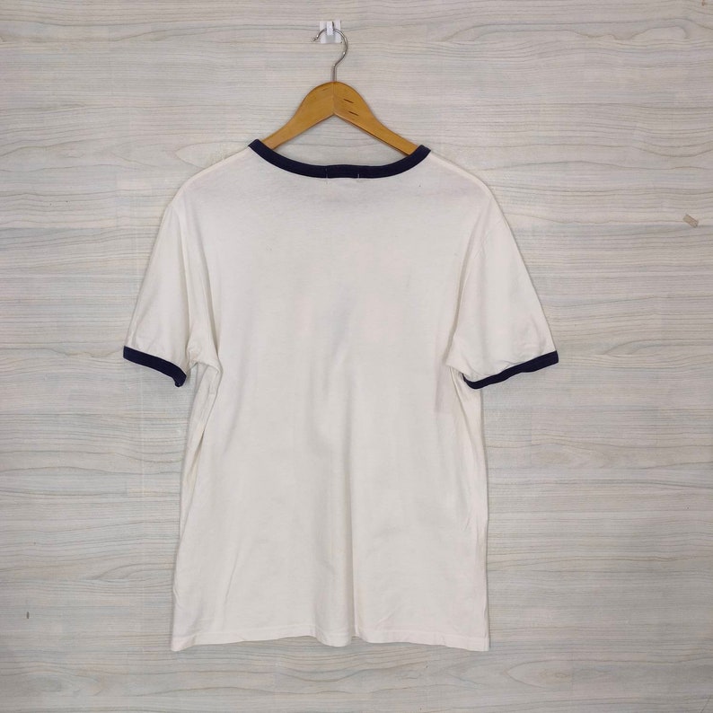 May include: White t-shirt with a navy blue ringer neckline and sleeve trim. The shirt is hanging on a wooden hanger against a woodgrain background.