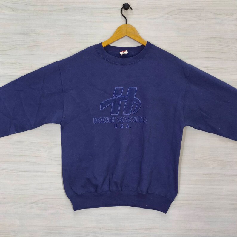 Pode incluir: Uma camisola azul-marinho com um logotipo bordado a azul que diz "North Carolina U.S.A."