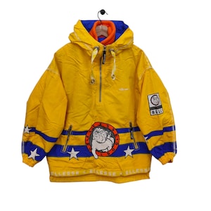Op de afbeelding: Gele vintage Ellessee anorakjas met blauwe en witte accenten. Voorzien van een bulldoglogo en sterdetails. Inclusief capuchon en een halve ritssluiting. Het woord "Ellesse" staat op de jas.
