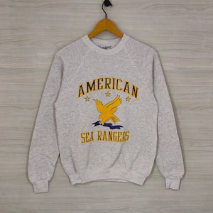May include: A grey sweatshirt with a yellow eagle graphic and the text "AMERICAN SEA RANGERS" printed on the front.