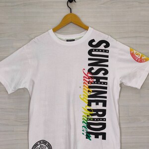 Puede incluir: Camiseta blanca con las palabras "SUNSHINE RIDE" en letras negras grandes en el lateral. La palabra "riding" est&aacute; en una escritura colorida. Un gr&aacute;fico circular est&aacute; en la parte inferior y un logotipo en la manga.