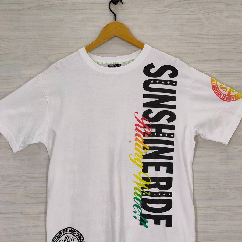 Puede incluir: Camiseta blanca con las palabras "SUNSHINE RIDE" en letras negras grandes en el lateral. La palabra "riding" est&aacute; en una escritura colorida. Un gr&aacute;fico circular est&aacute; en la parte inferior y un logotipo en la manga.