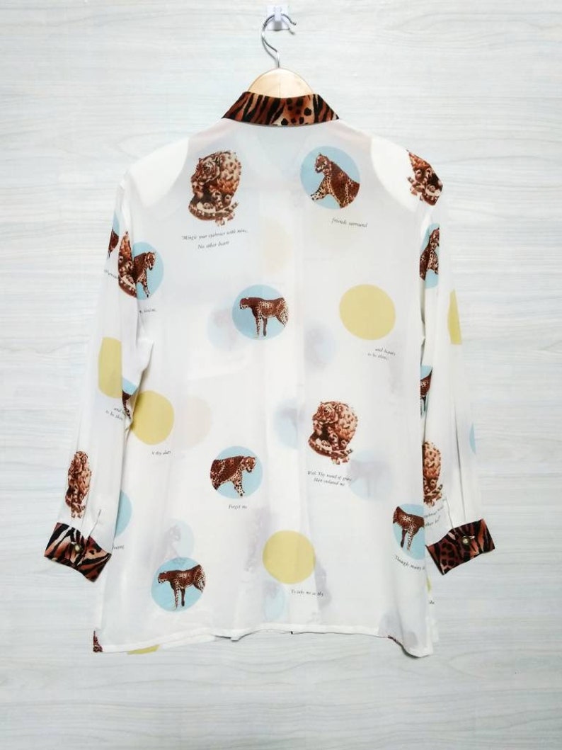 May include: White long-sleeved button-up shirt with a leopard print collar and cuffs. The shirt features a white background with a repeating pattern of circles containing leopard print images and text. The text reads: "Wings were given to us, not other burdens." "Fenced vineyard." "And beauty is in the eye." "What the world of pain has meant to me." "Though many waters cannot quench love." "It is a day in July." "Forget me not."