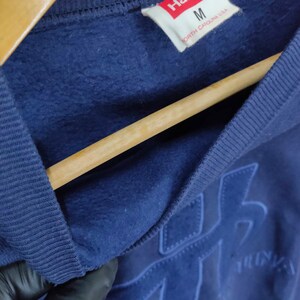 Pode incluir: Uma camisa de moletom azul marinho com uma etiqueta branca que diz "Hanes" e "M" para o tamanho. A etiqueta tamb&eacute;m diz "Carolina do Norte, EUA". A camisa de moletom tem um grande logotipo bordado na frente.