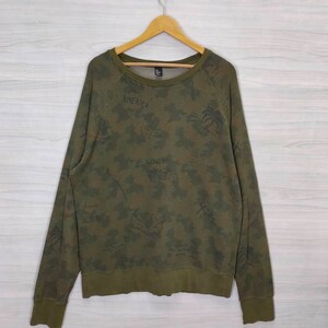Könnte beinhalten: Grüner Camouflage-Sweatshirt mit einem grafischen Druck von Palmen, einem Motorrad und dem Wort "ROCK" in Schwarz. Der Sweatshirt hat auch das Wort "AMERICA" auf der Vorderseite gedruckt.