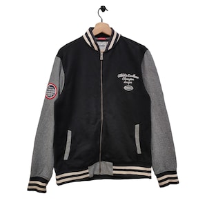Puede incluir: Chaqueta de béisbol negra y gris con rayas blancas en los puños y en la parte inferior. La chaqueta tiene una cremallera y un parche en la manga izquierda que dice "Champion League". La parte posterior de la chaqueta tiene el texto "Athletic Excellence Champion League" en blanco.