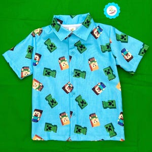 May include: A light blue short-sleeved button-down shirt with a repeating pattern of green pixelated characters and brown pixelated characters.