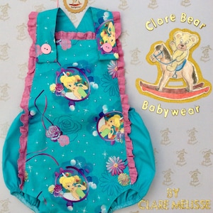 May include: A turquoise baby romper with pink ruffles and a Tinkerbell print. The romper has a pink button on the front and a white tag that says "Clare Bear Babywear".