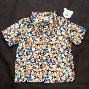 May include: A white short-sleeved button-down shirt with a repeating pattern of Mickey Mouse, Donald Duck, and Goofy characters.