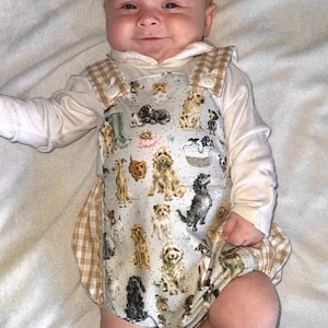 May include: A baby wearing a light blue romper with a pattern of various dog breeds. The romper has a white long-sleeved shirt underneath and tan and white checkered accents. The baby is lying on a white surface.